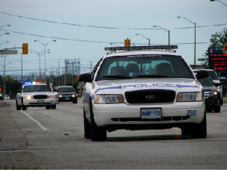 A photograph shows two police cars driving, one with its lights flashing.