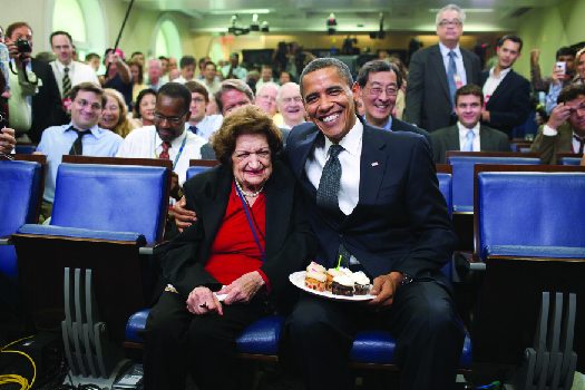 An image of Barack Obama and Helen Thomas seated. Obama holds a plate of cake.