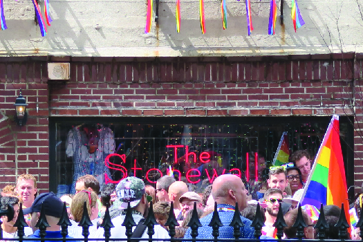 An image of a group of people standing in front of a brick building. A sign in the window of the building reads “The Stonewall”. A multicolored flag is held by a person to the right.