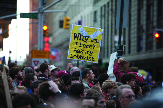 An image of a crowd of people, one of whom holds a sign that reads “Why am I here? Ask my lobbyist”.